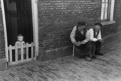 221132 Afbeelding van twee oudere mannen op een krukje en een kind achter een hekje in de deuropening van een huis aan ...